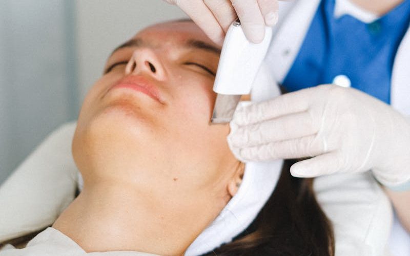 Crop unrecognizable female cosmetologist in medical uniform and gloves doing ultrasonic face cleaning procedure in beauty clinic